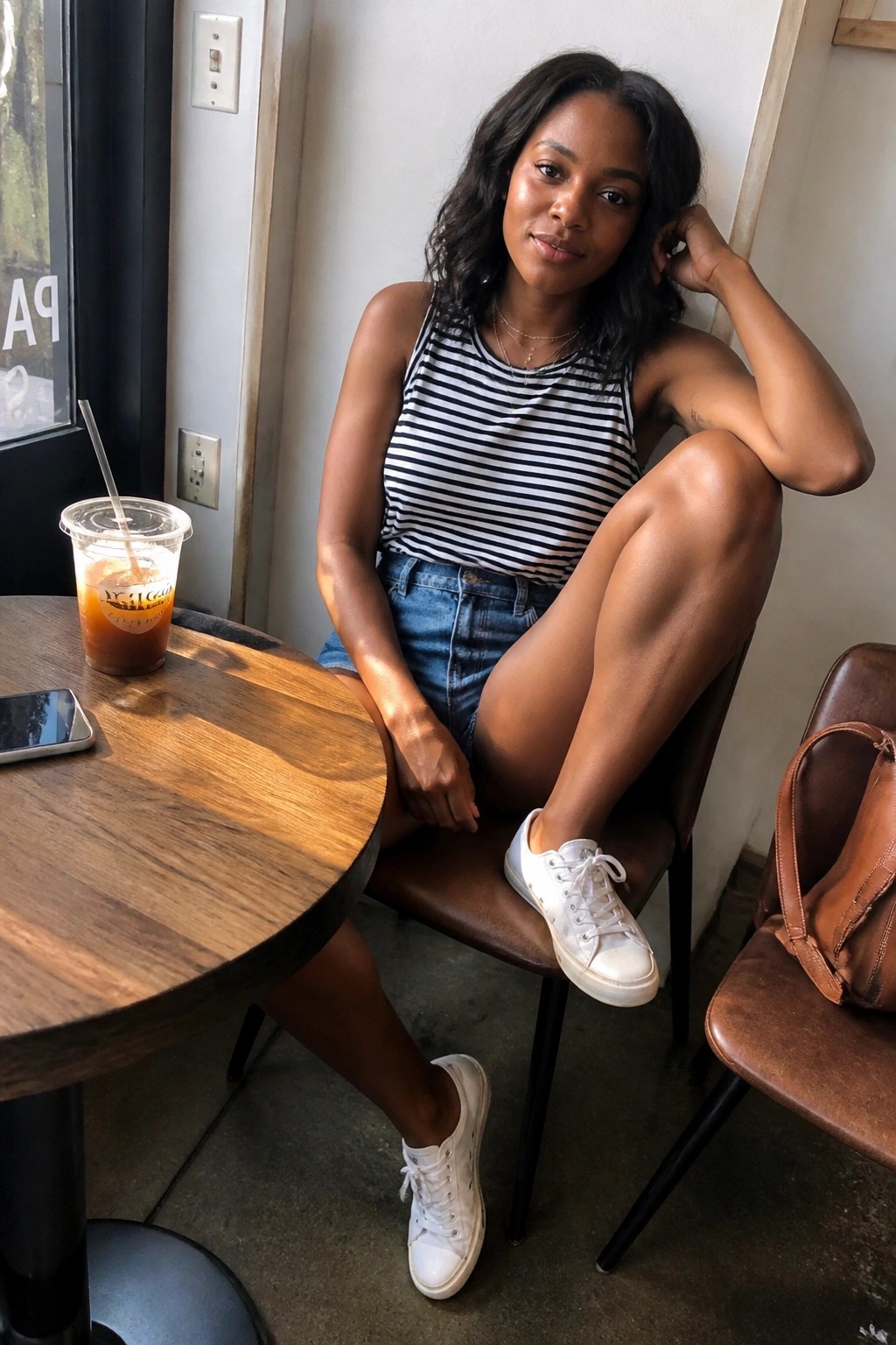 Woman in striped tank top and denim shorts at café table with iced coffee