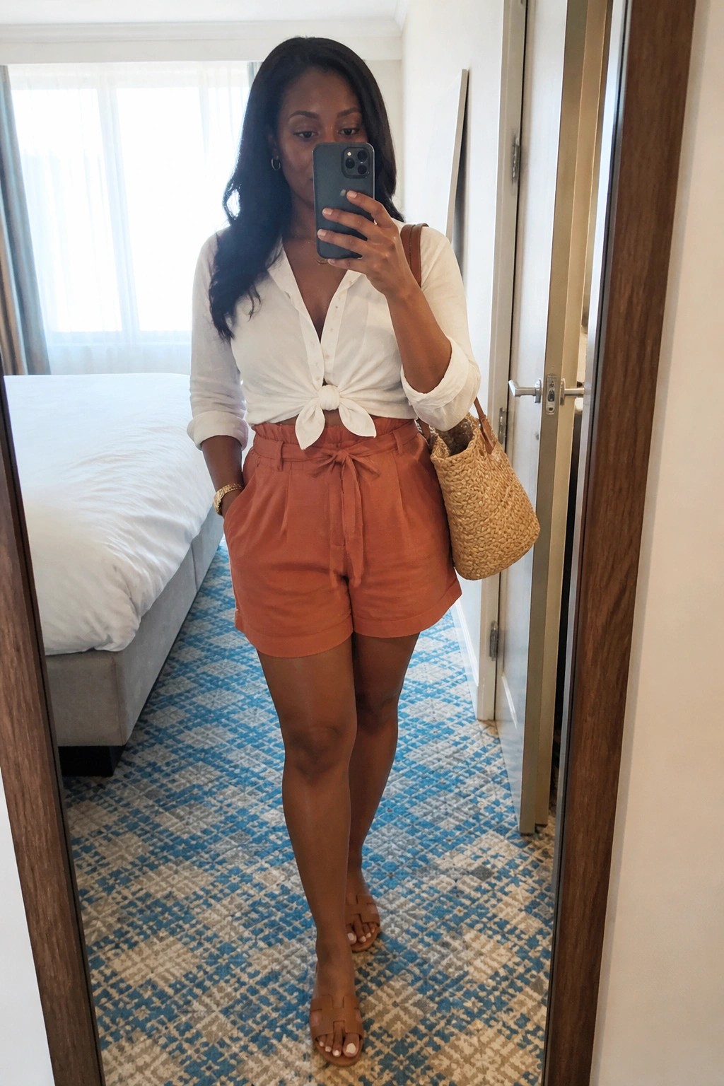 Woman wearing a white button-down shirt tied at the waist with orange high-waisted shorts, carrying a straw bag and brown slide sandals while taking a mirror selfie.