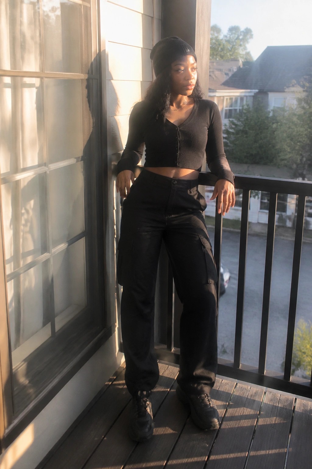 A woman in an all-black outfit featuring a cropped ribbed cardigan, high-waisted cargo pants, chunky boots, and a beanie with layered necklaces.