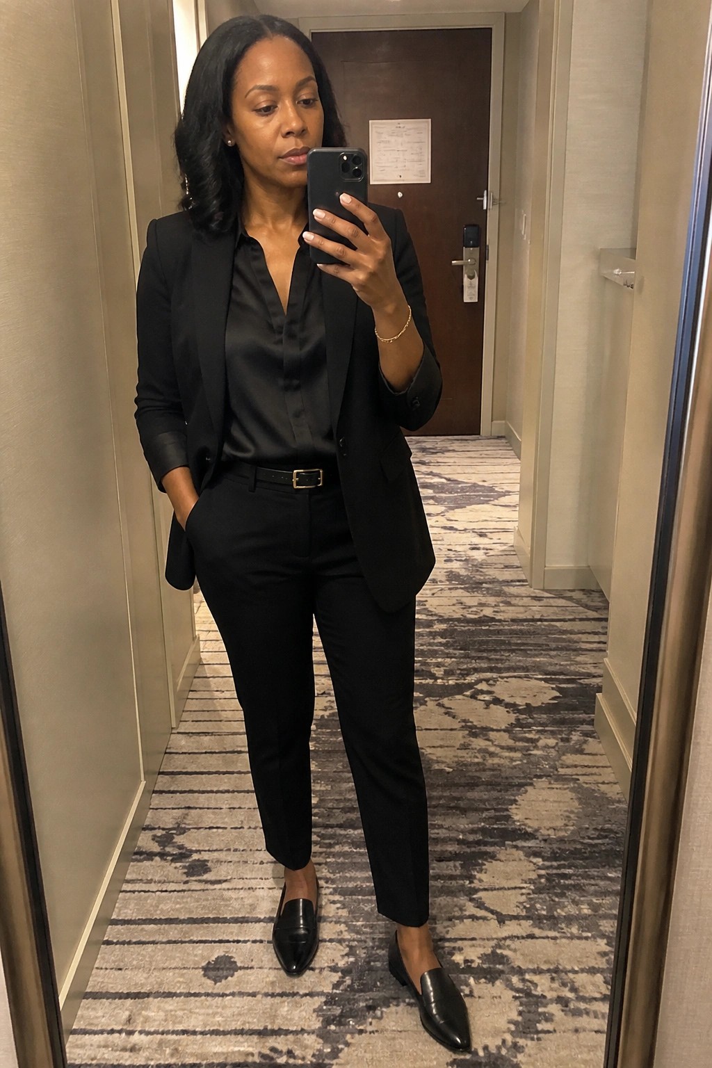 Woman in black suit taking mirror selfie in hotel hallway with patterned carpet.