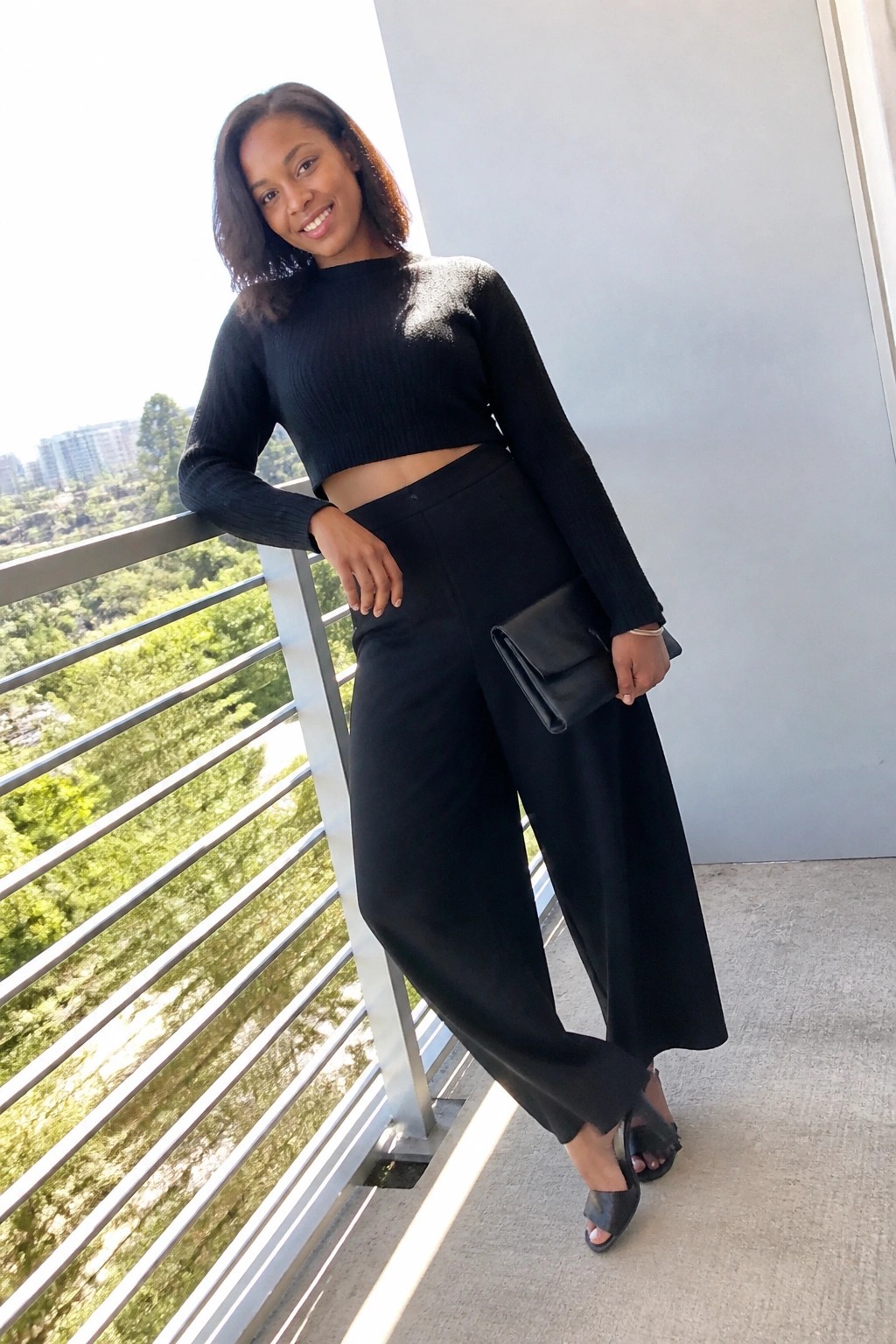 Smiling woman in black crop top and wide-leg pants on balcony holding clutch