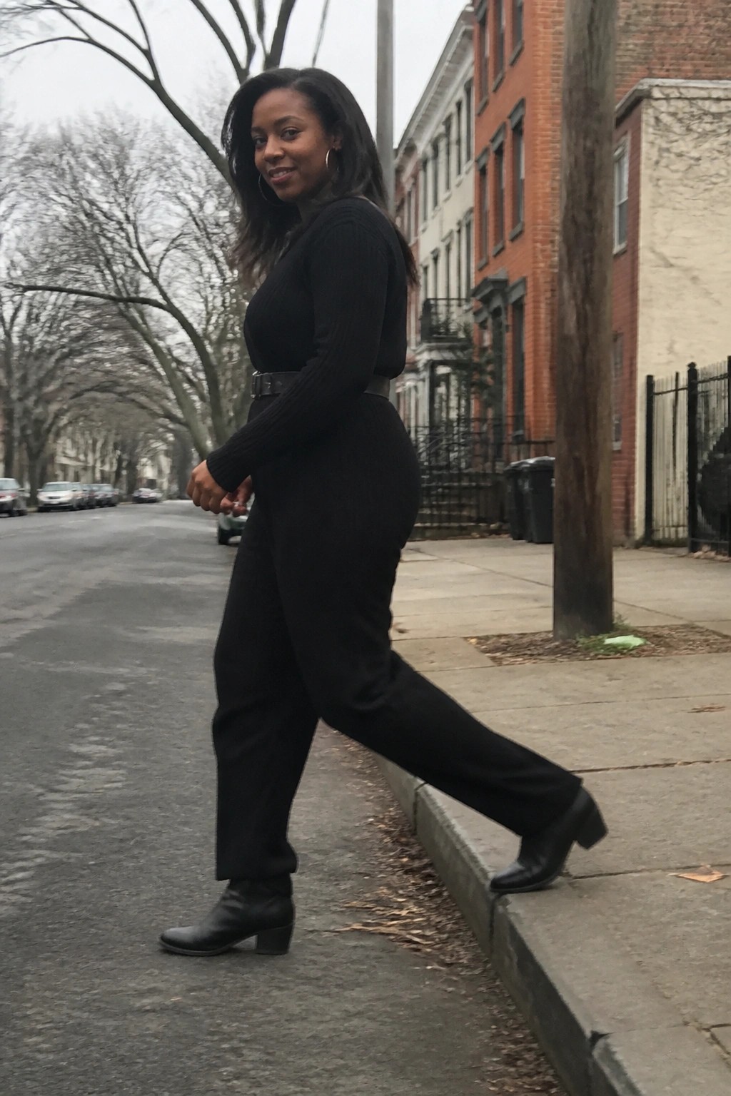 Woman in black jumpsuit posing with leg raised on urban sidewalk