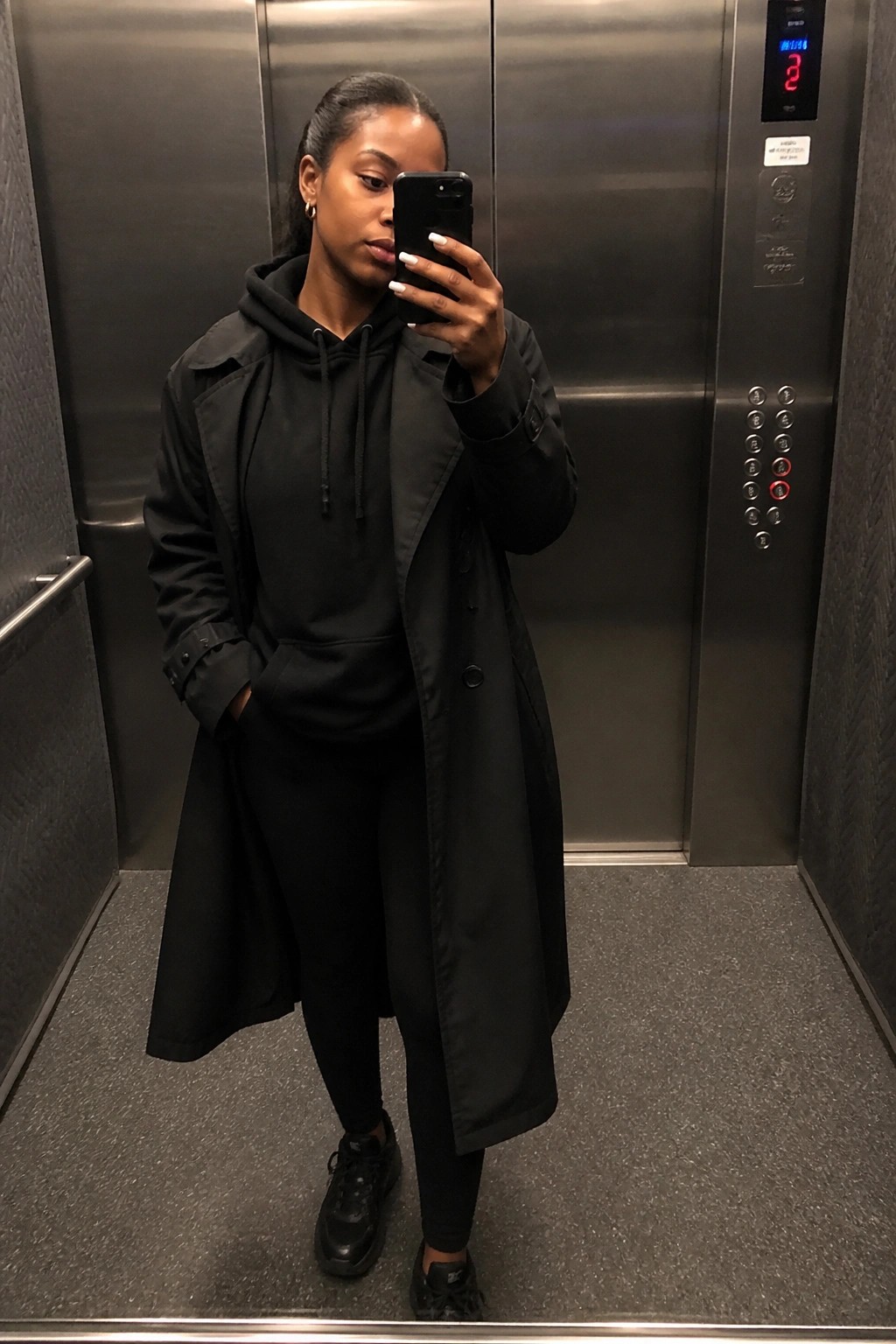 A woman in an all black outfit consisting of a long trench coat over a hoodie paired with leggings and sneakers taking a mirror selfie inside an elevator.