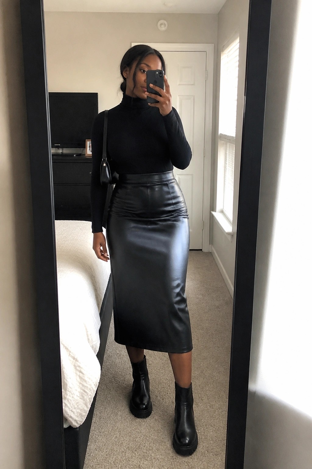 Woman in black turtleneck and leather midi skirt taking bedroom mirror selfie