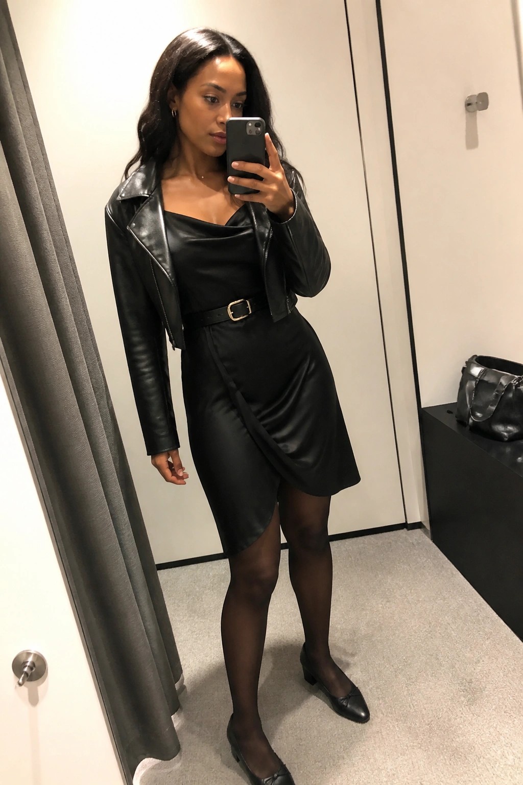 Woman in black leather jacket and dress taking mirror selfie in fitting room