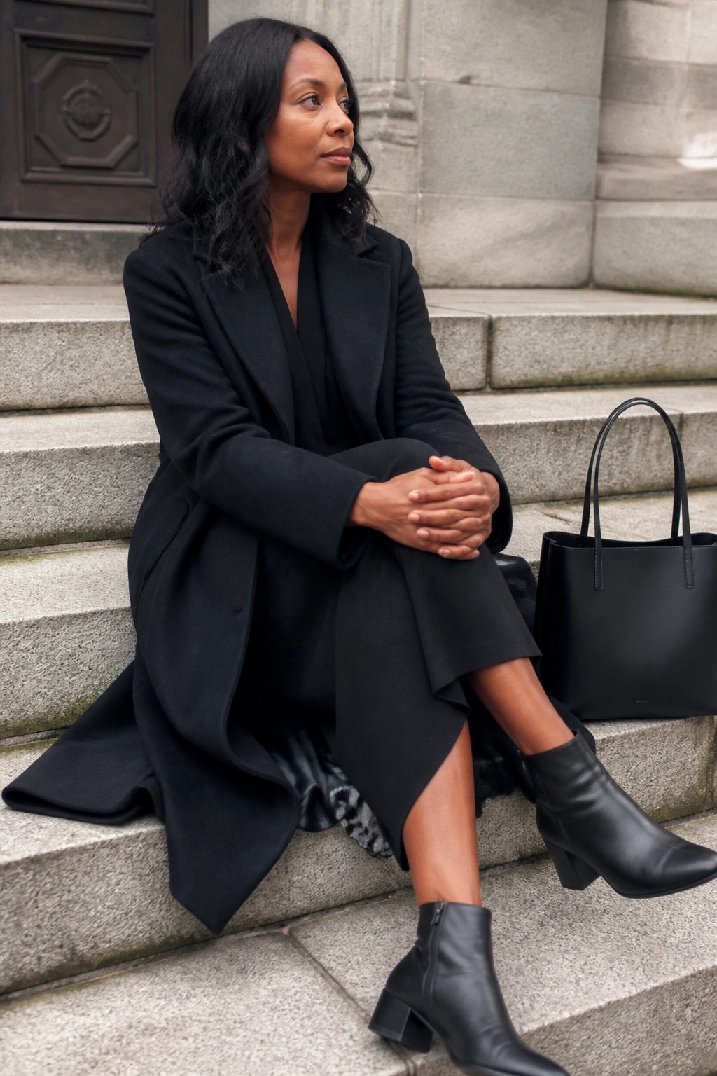 Woman in black coat and boots seated on stone steps beside handbag