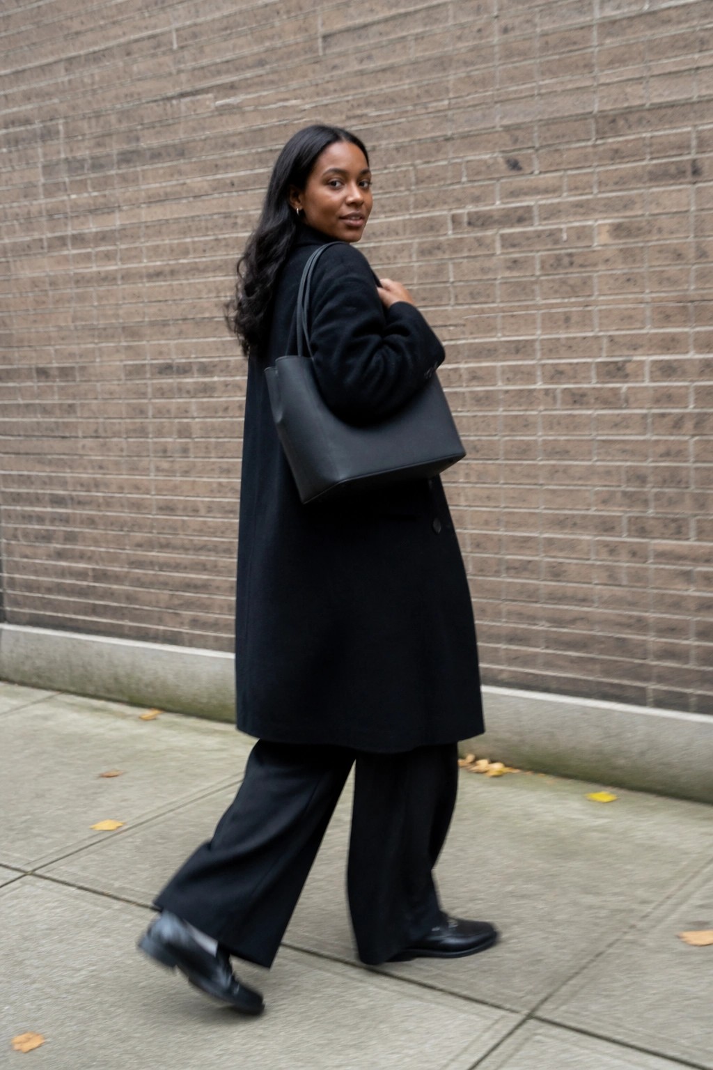 Woman in black coat with tote bag walks past brick wall, glancing back