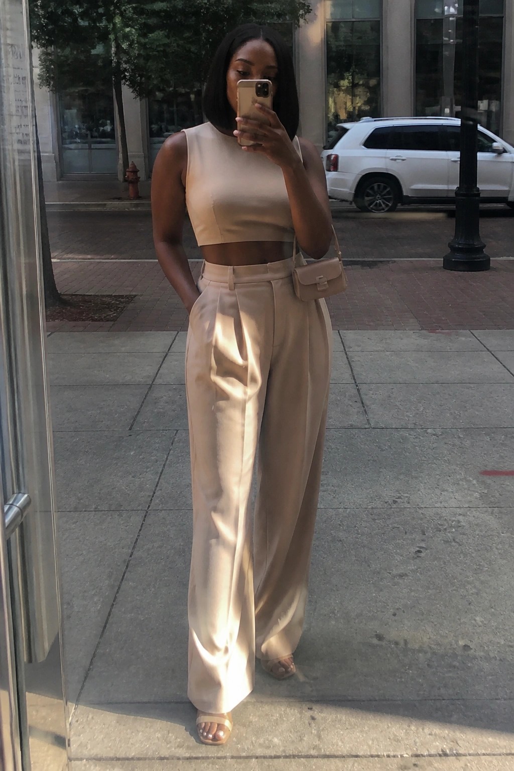 Woman in beige crop top and wide-leg pants taking selfie on city sidewalk