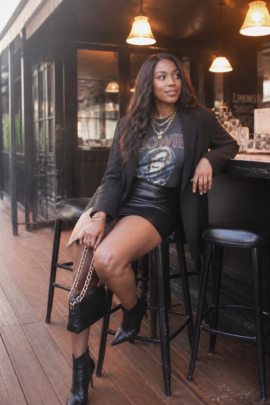 Woman wearing black blazer over gray graphic Bowie t-shirt, black leather mini skirt, black ankle boots, and black handbag with gold chain strap, accessorized with layered gold necklaces and bracelets.
