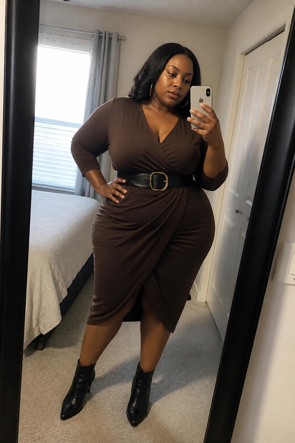 Plus size woman wearing a chocolate brown wrap midi dress with a black belt and gold buckle paired with black ankle boots while taking a mirror selfie in a bedroom.