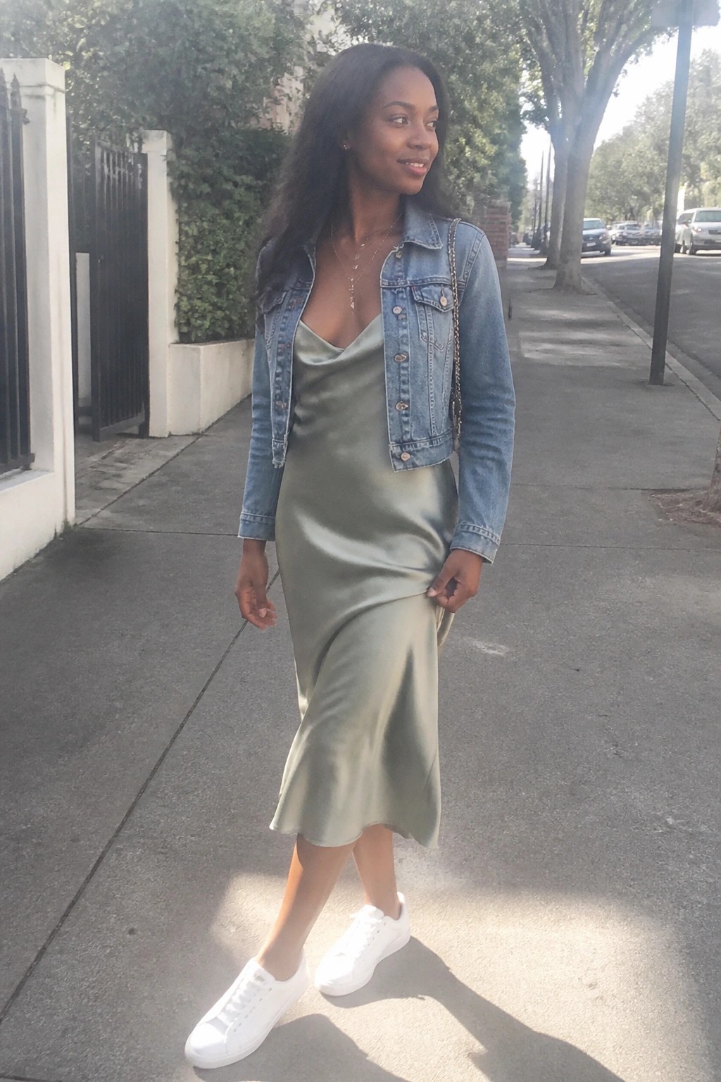 Woman in sage green satin slip dress with cropped denim jacket on sunny sidewalk