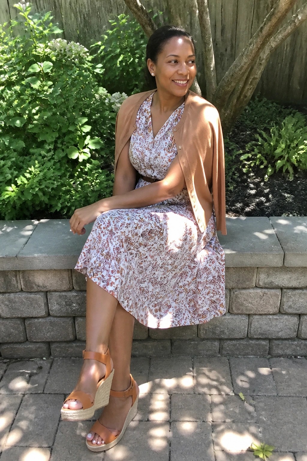 A Black woman wearing a white floral midi dress with brown and rust prints layered under an open brown cardigan and paired with matching brown wedge sandals.