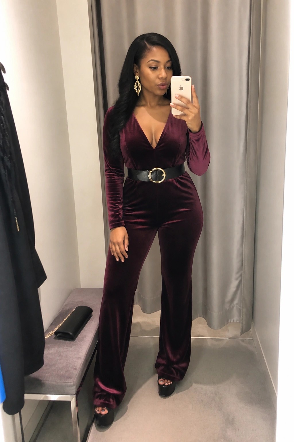 Woman in burgundy velvet jumpsuit taking mirror selfie in fitting room.