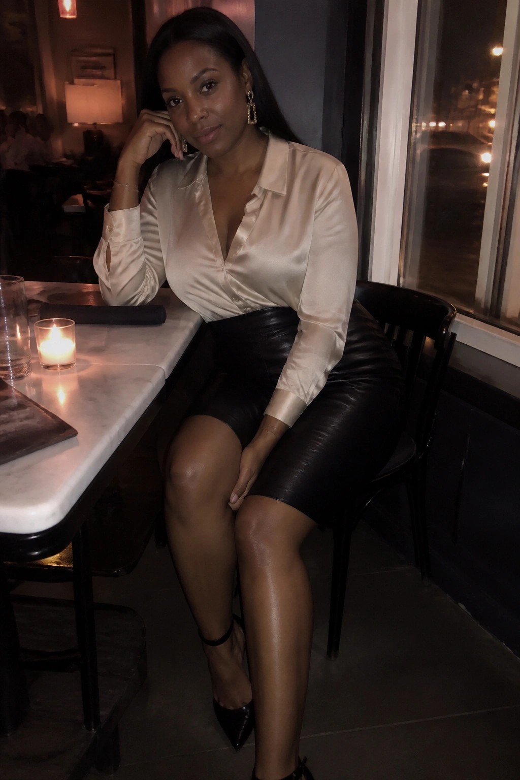 Woman wearing a cream satin long sleeve button up blouse tucked into a black leather mini skirt, paired with black pointed toe heels and gold jewelry.
