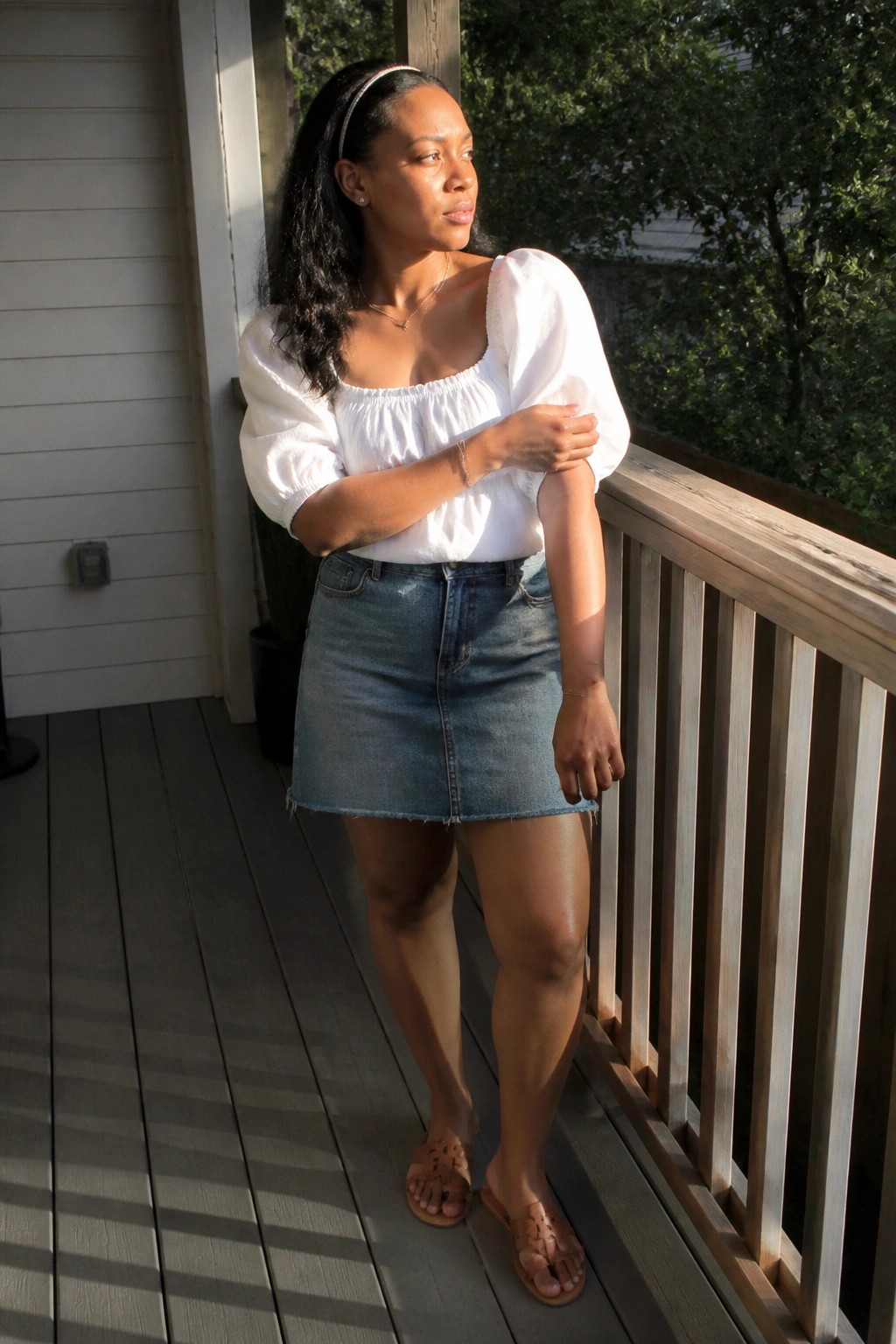 Woman in white off-shoulder top and denim skirt on sunny wooden porch