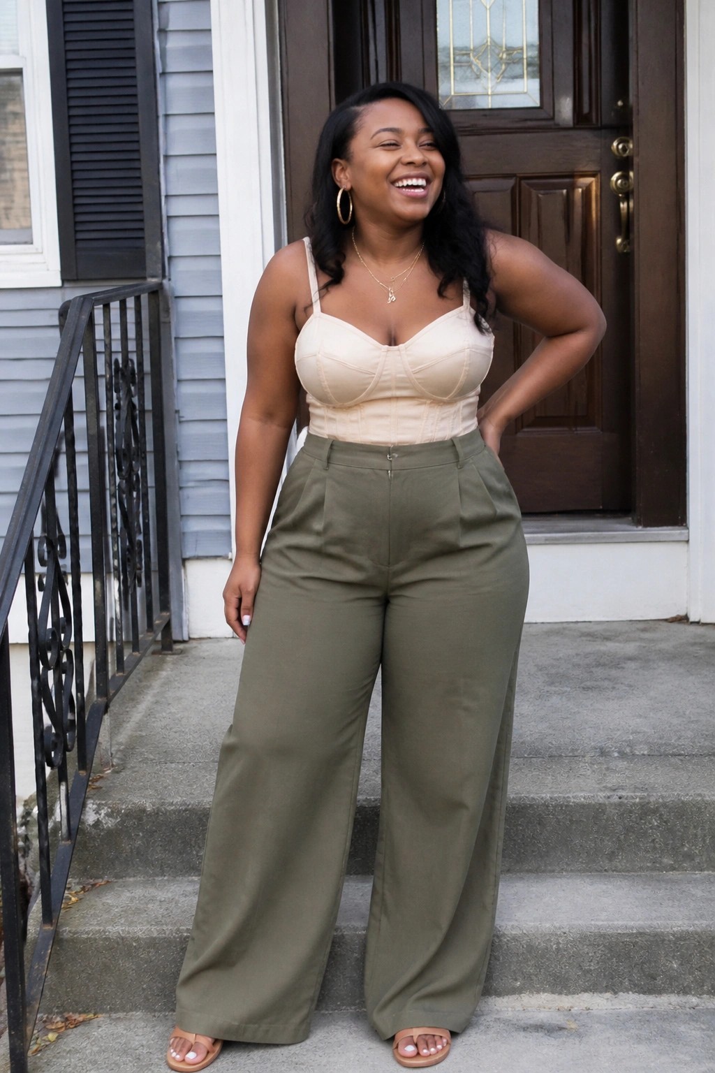 Smiling woman in beige corset top and olive wide-leg pants on steps.