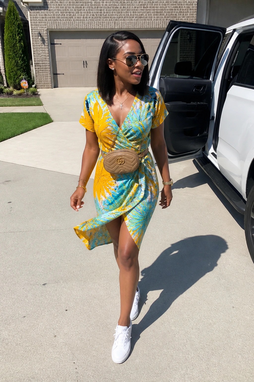 Woman in yellow-turquoise floral wrap dress and white sneakers by open SUV door.