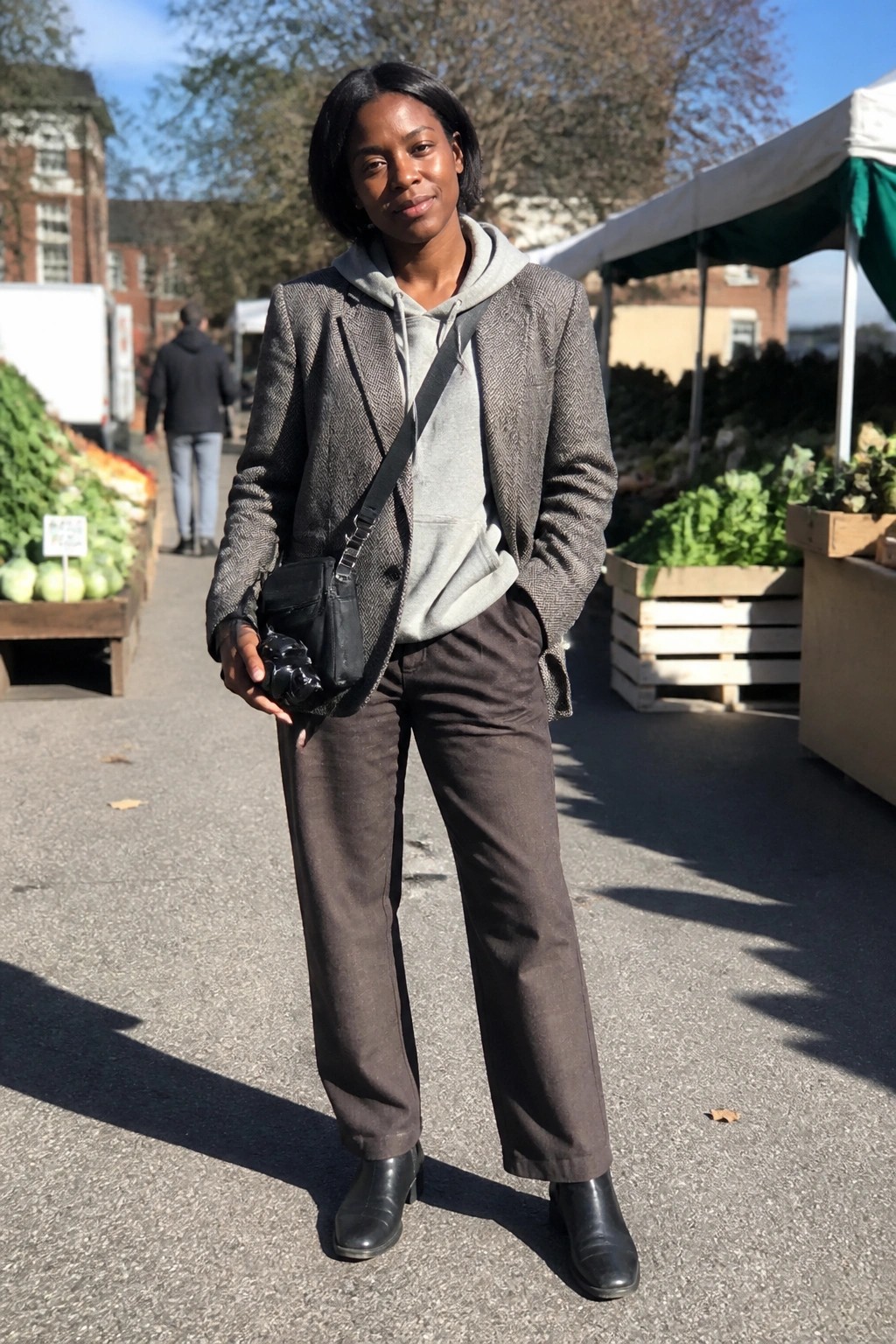 Woman in herringbone blazer over grey hoodie, brown straight trousers and black ankle boots holding a small black bag at an outdoor market.