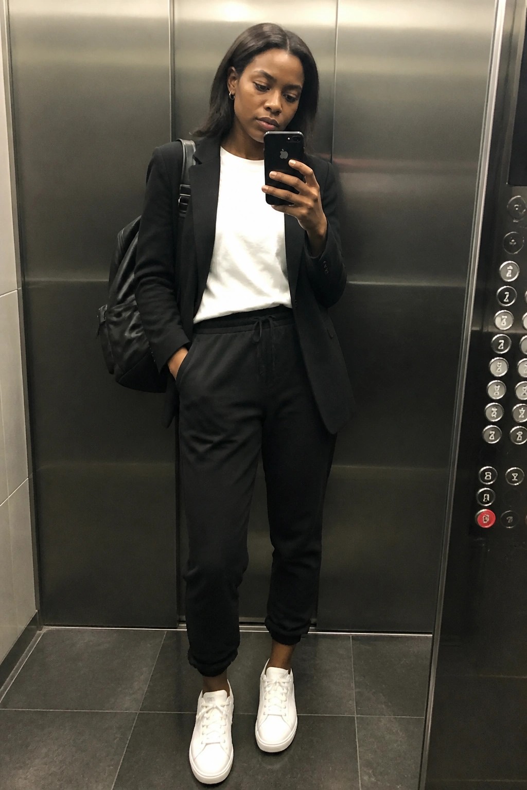 Woman in black blazer, white tee, and joggers holding phone in elevator