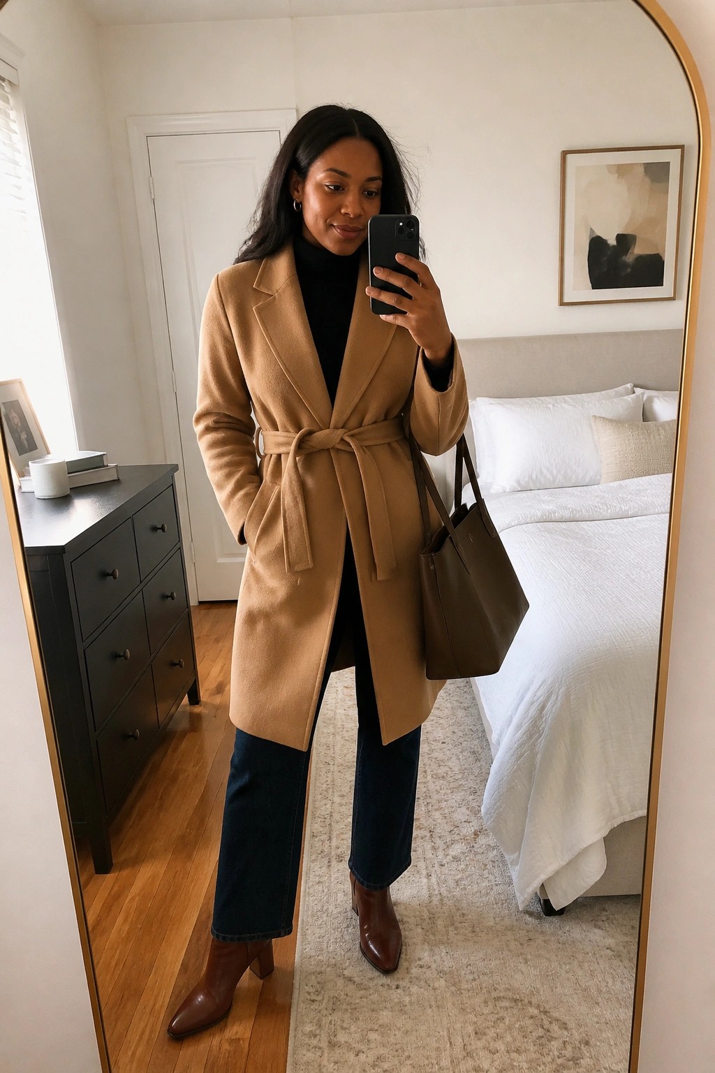 Woman in tan belted coat holding phone and bag in bedroom mirror