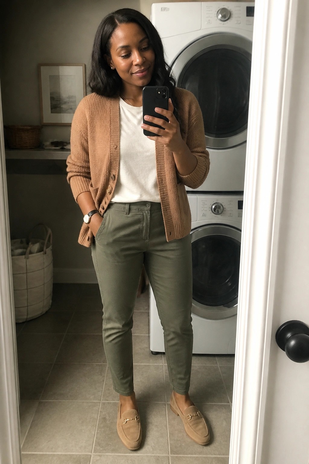 Woman in tan cardigan and olive pants taking mirror selfie in laundry room