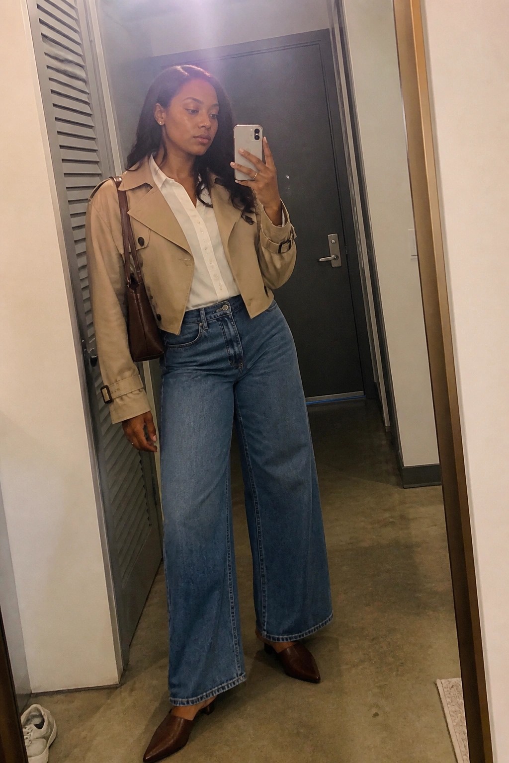 Woman in beige cropped trench jacket and wide-leg jeans taking mirror selfie with phone