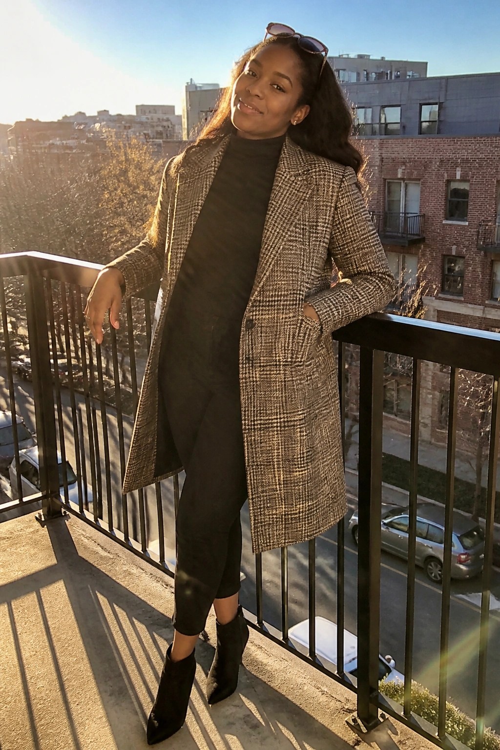 Woman in brown plaid coat posing on sunny urban balcony with city view