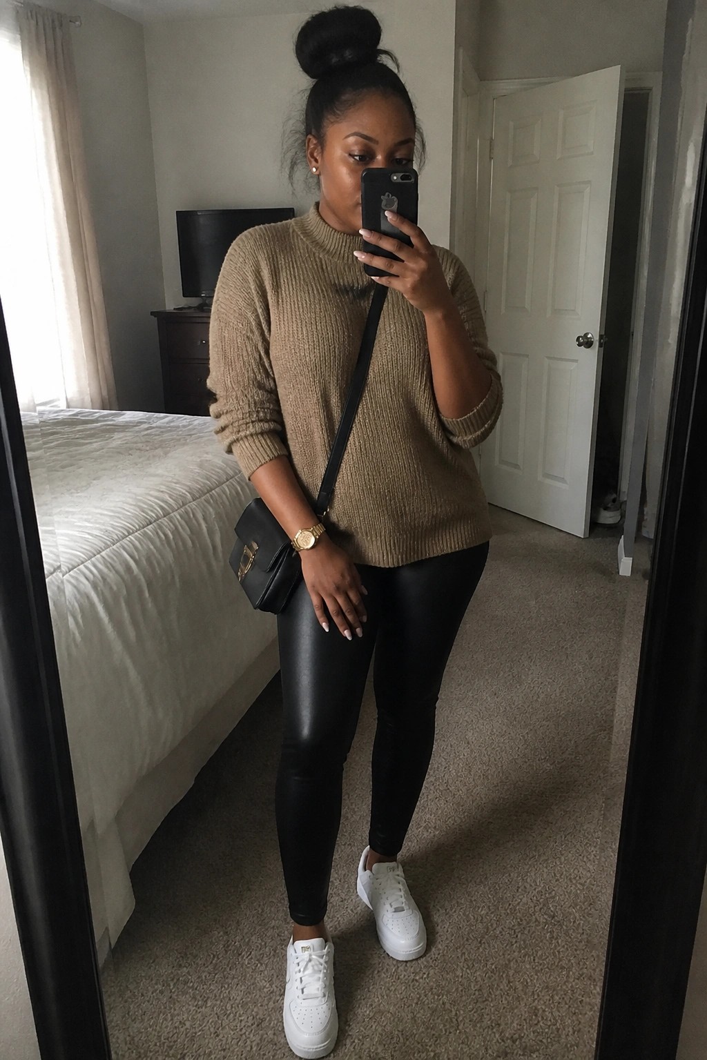 Woman in tan sweater and black leggings taking bedroom mirror selfie