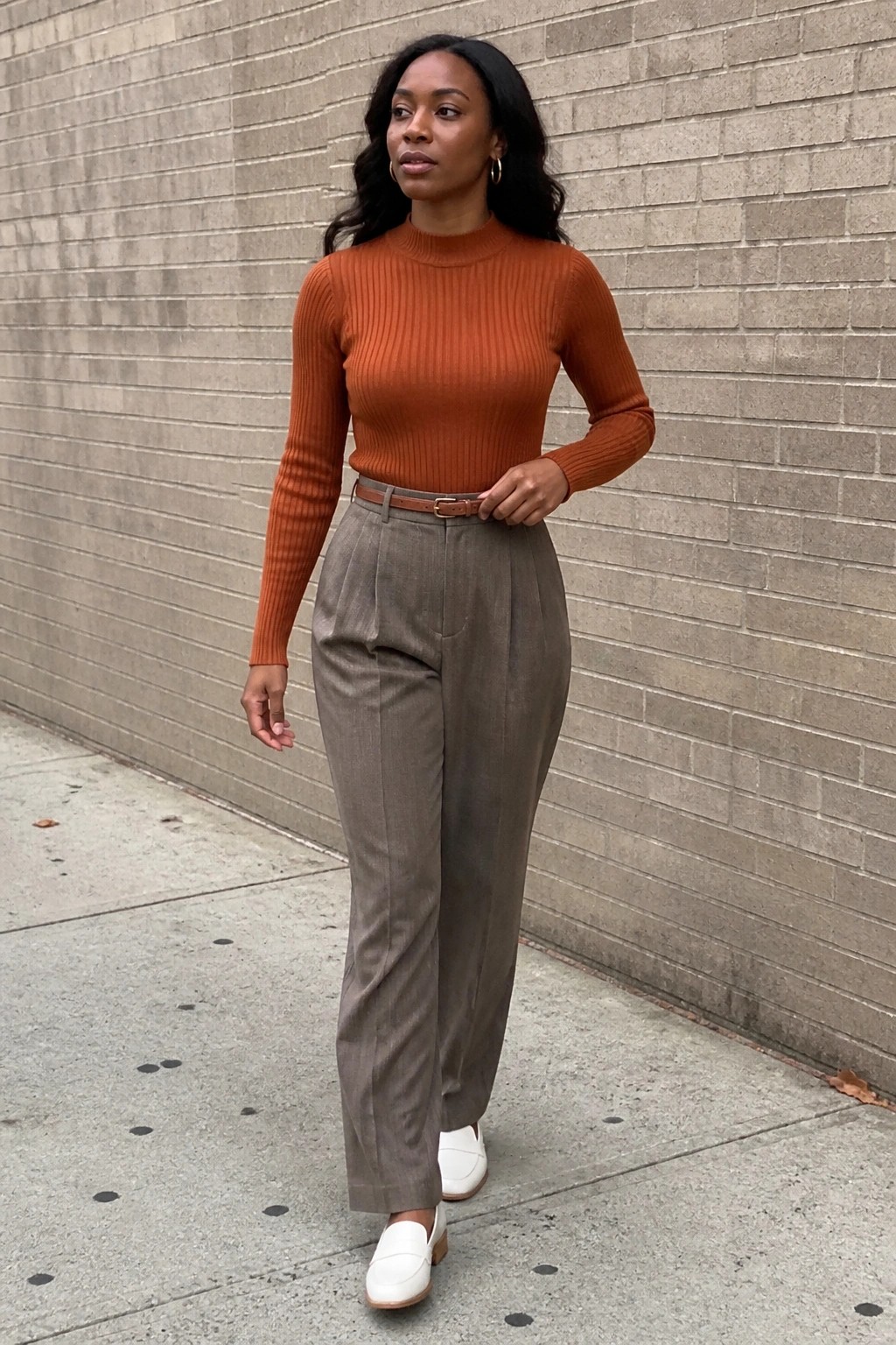 Woman in orange turtleneck, gray pleated trousers, and white loafers by brick wall.