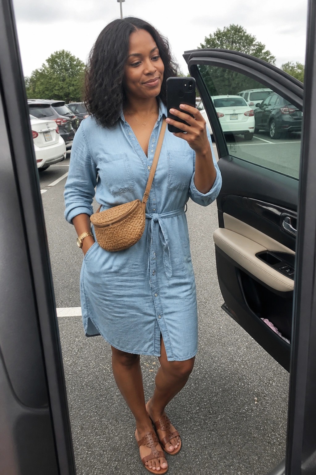 Woman in light blue denim shirt dress taking selfie by open car door in lot