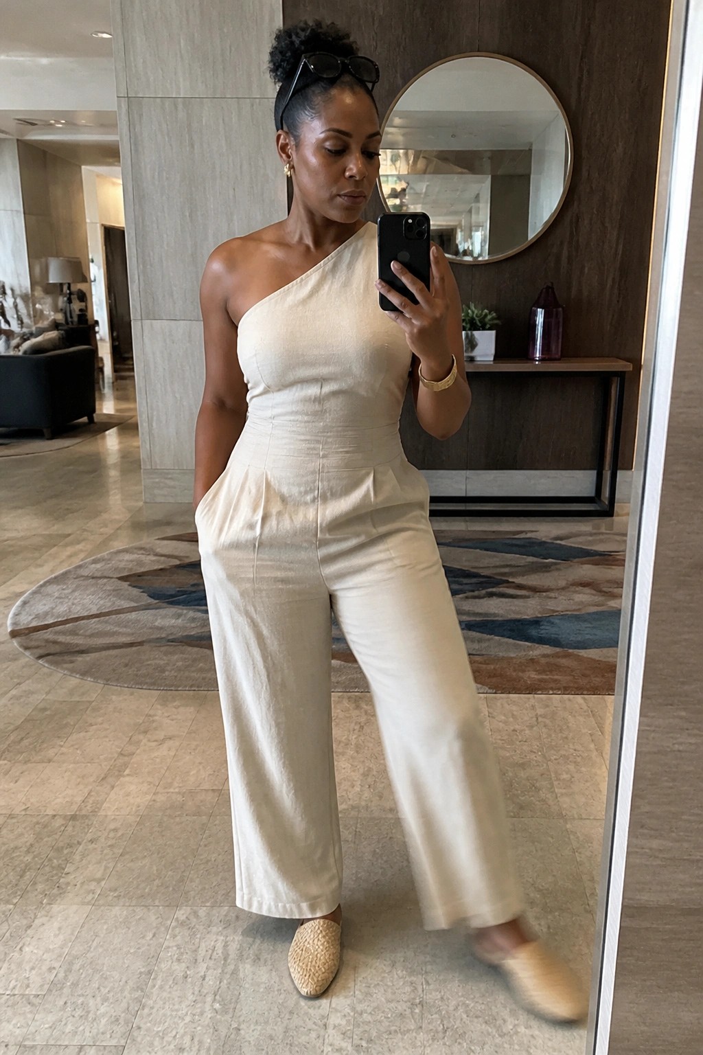 Woman in cream one-shoulder jumpsuit taking mirror selfie in elegant lobby