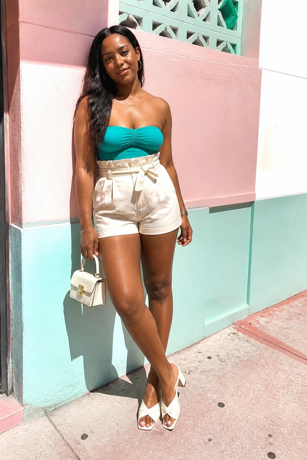 Woman in teal strapless top and white belted shorts holding handbag by colorful wall