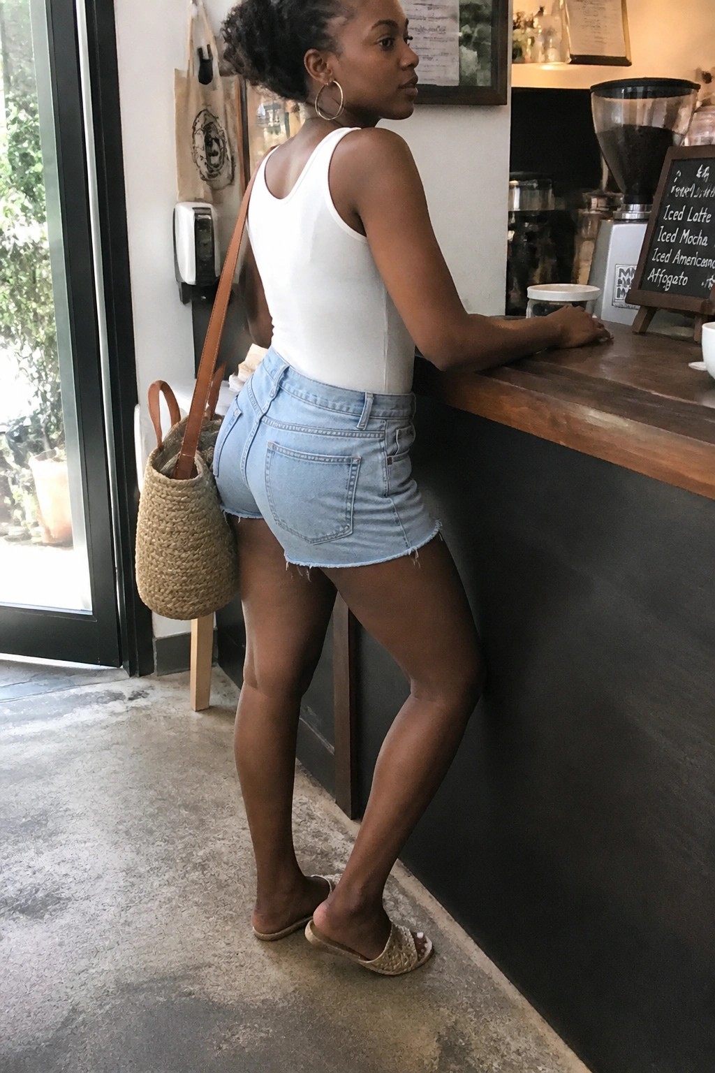Woman in white tank top and denim shorts leaning on cafe counter with woven bag