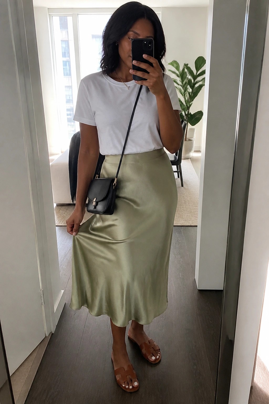 Woman in white tee and sage green satin skirt taking mirror selfie with black bag