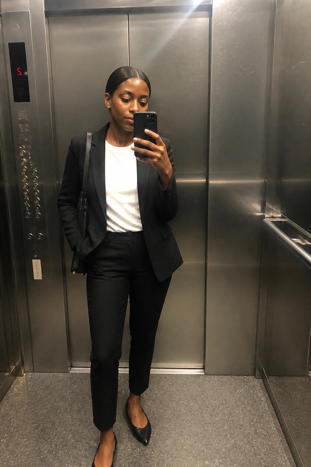Woman in black blazer and pants taking selfie inside metallic elevator.