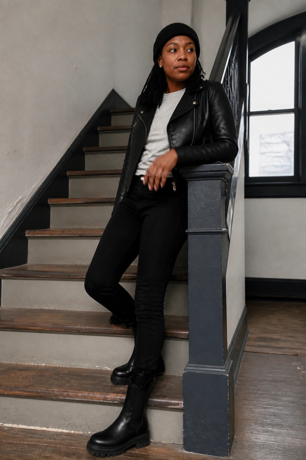 Woman in black leather jacket and beanie leaning on wooden staircase railing