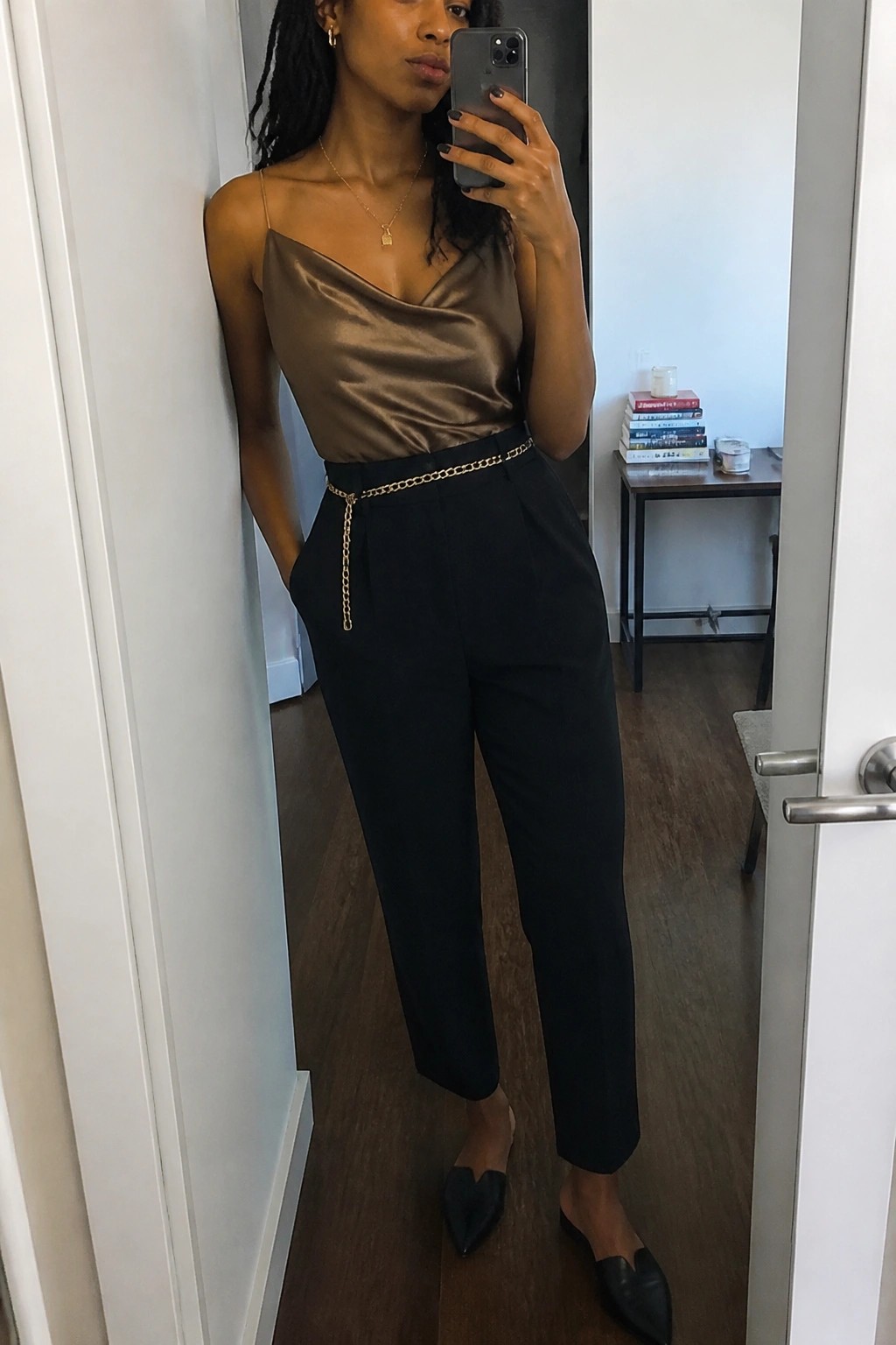 Woman in brown satin top and black pants with gold chain belt taking mirror selfie