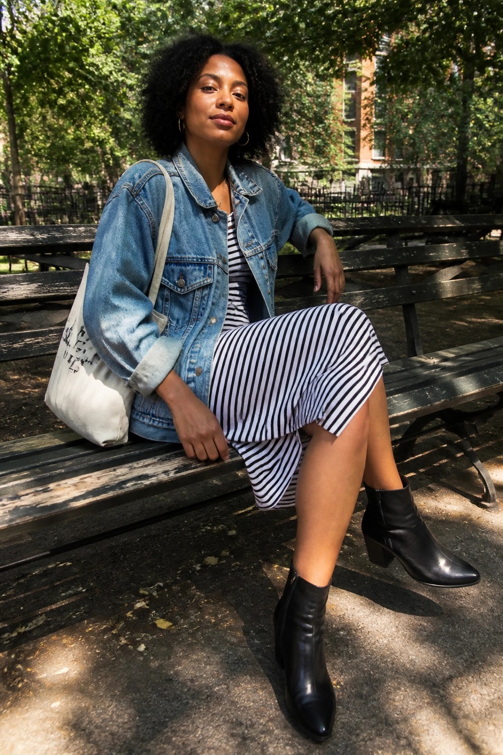 Woman in denim jacket and striped dress on park bench with white tote bag