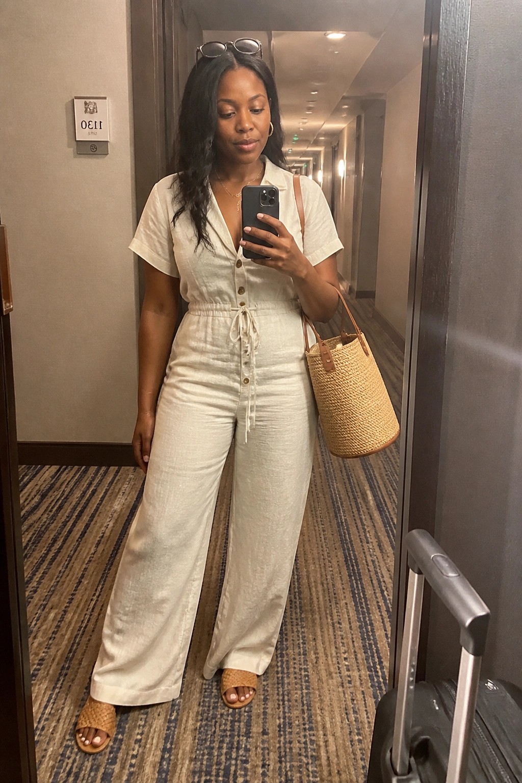 Woman in cream jumpsuit takes mirror selfie in hotel hallway with straw bag and suitcase