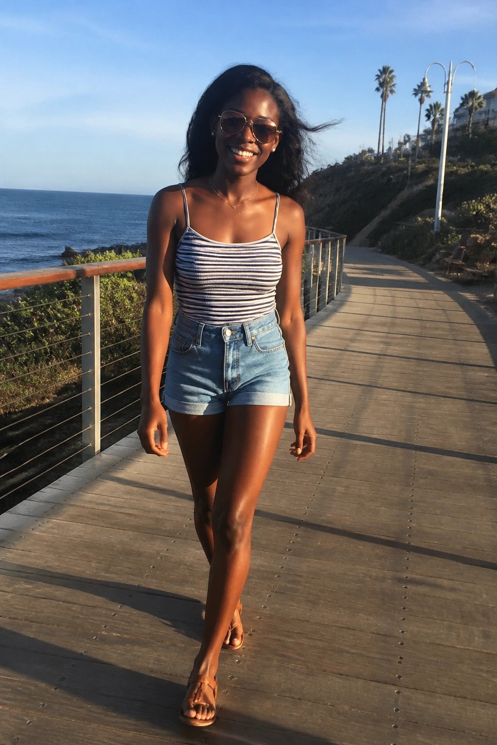Smiling woman in striped top and denim shorts on sunny ocean boardwalk