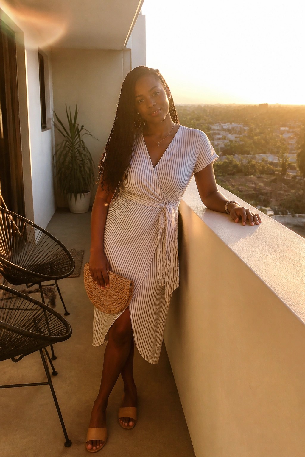 Woman wearing a white and navy striped wrap dress tied at the waist with short sleeves and a front slit, paired with tan slide sandals and a small woven clutch while standing on a balcony.