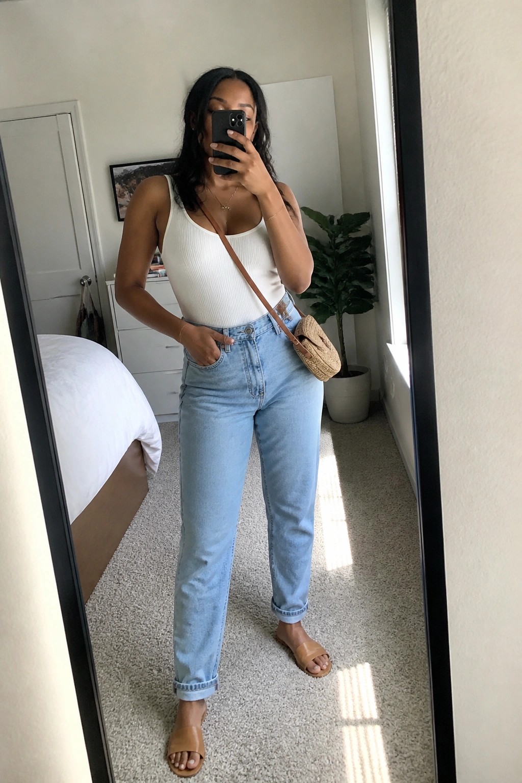 Woman in white tank top and light blue jeans taking mirror selfie in bedroom