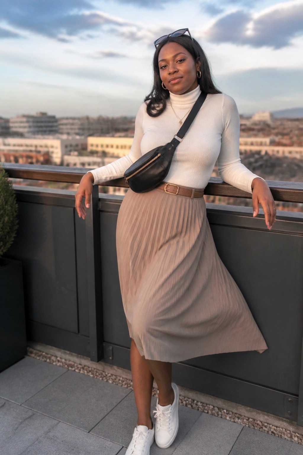 Woman in white turtleneck and beige pleated skirt with black bag on city balcony