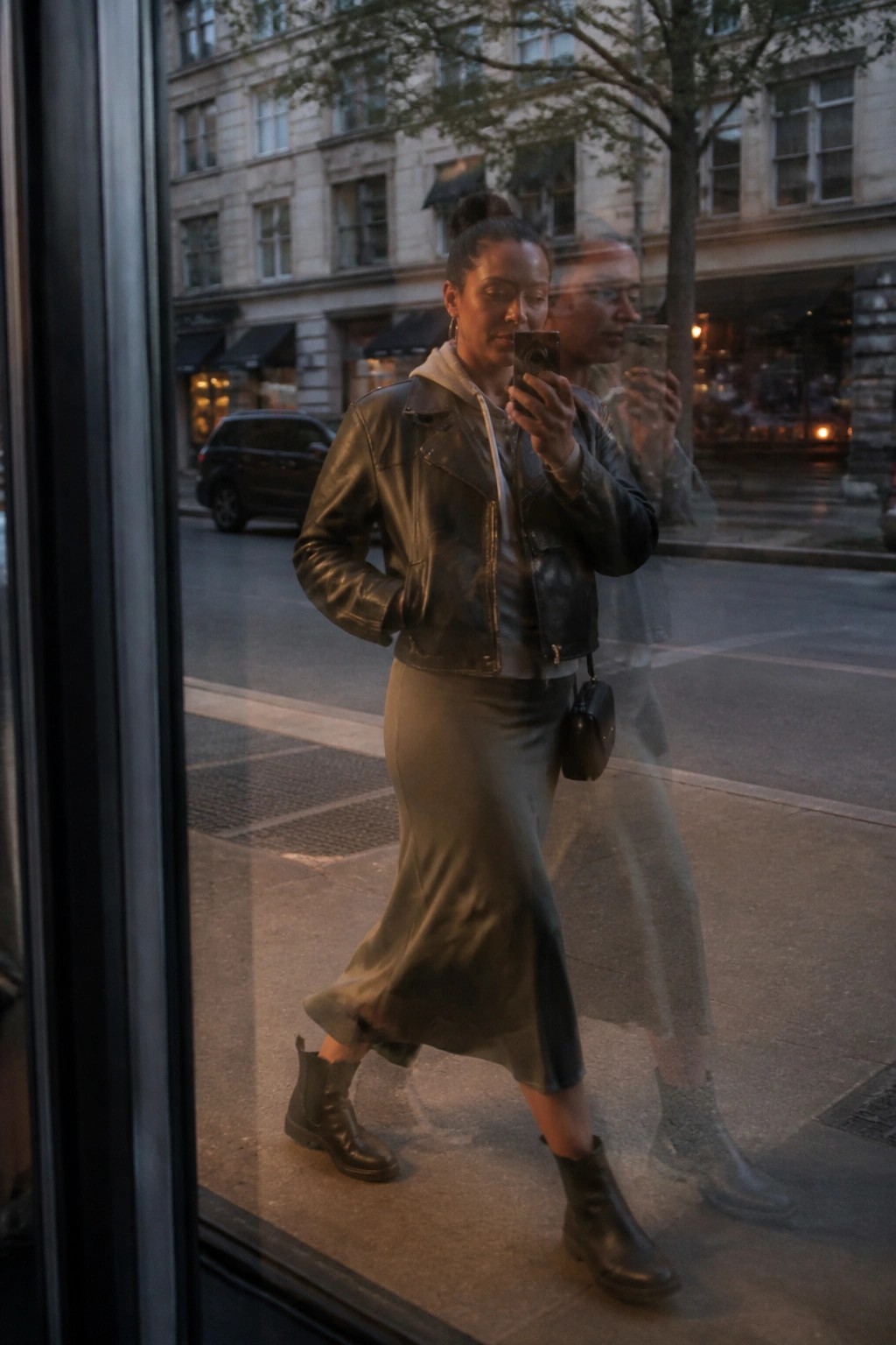 Woman wearing black leather jacket over grey hoodie, olive green satin midi skirt, and black ankle boots while holding a phone in a mirror reflection.