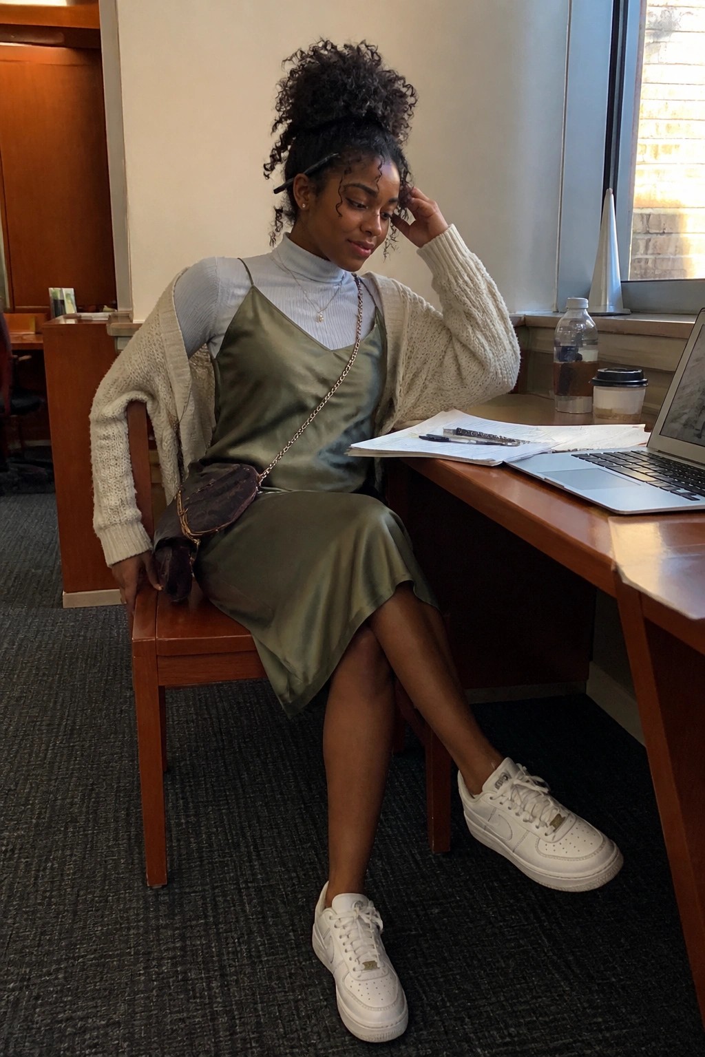 Woman in olive satin dress and cream cardigan seated at wooden study desk with laptop