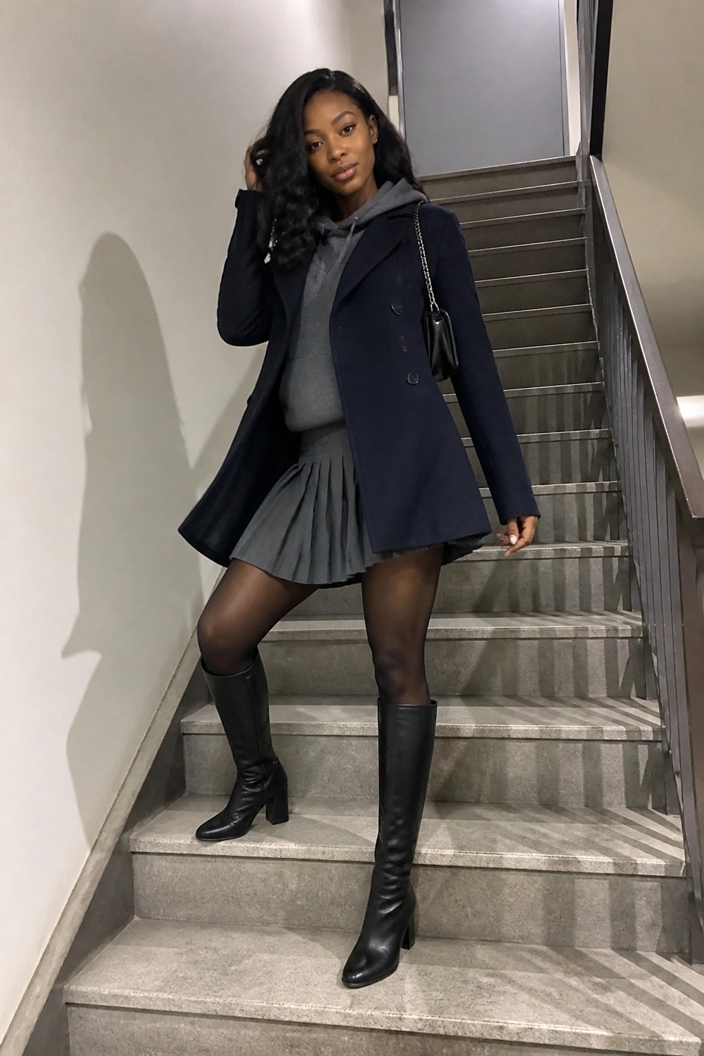 Black woman wearing a navy double breasted coat over a grey hoodie and matching pleated mini skirt with black tights and knee high boots while holding a small black chain strap bag.