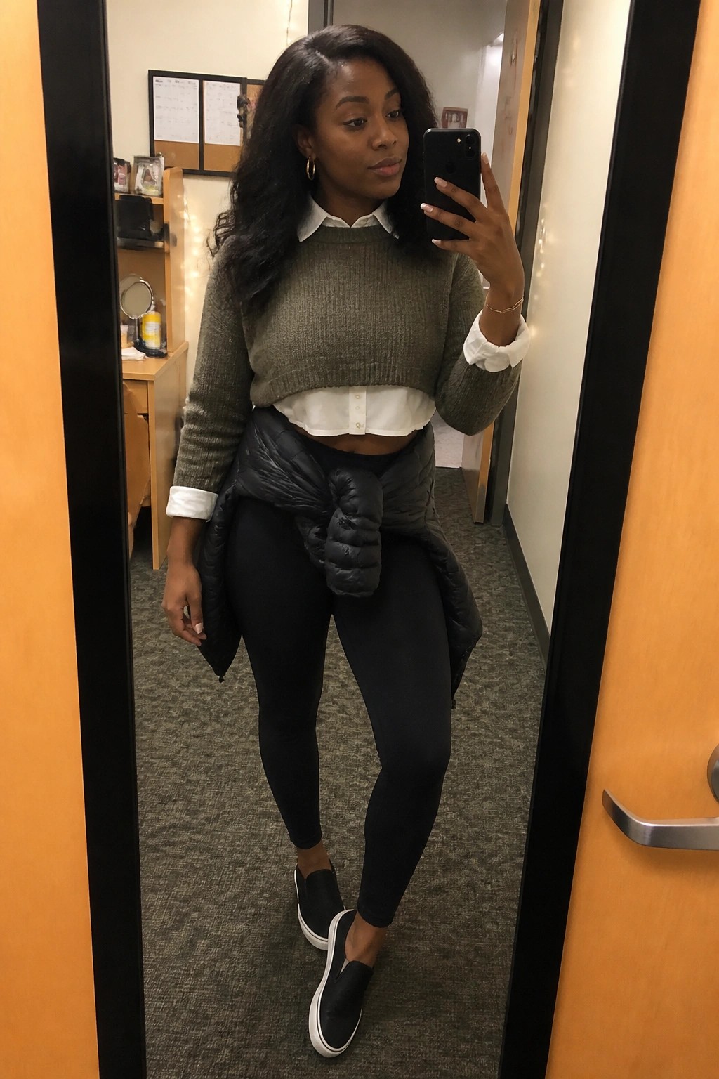 Woman in mirror selfie wearing an olive green cropped knit sweater over a white button down shirt with collar and cuffs visible, black puffy vest tied at the waist, black leggings, and black slip on sneakers.