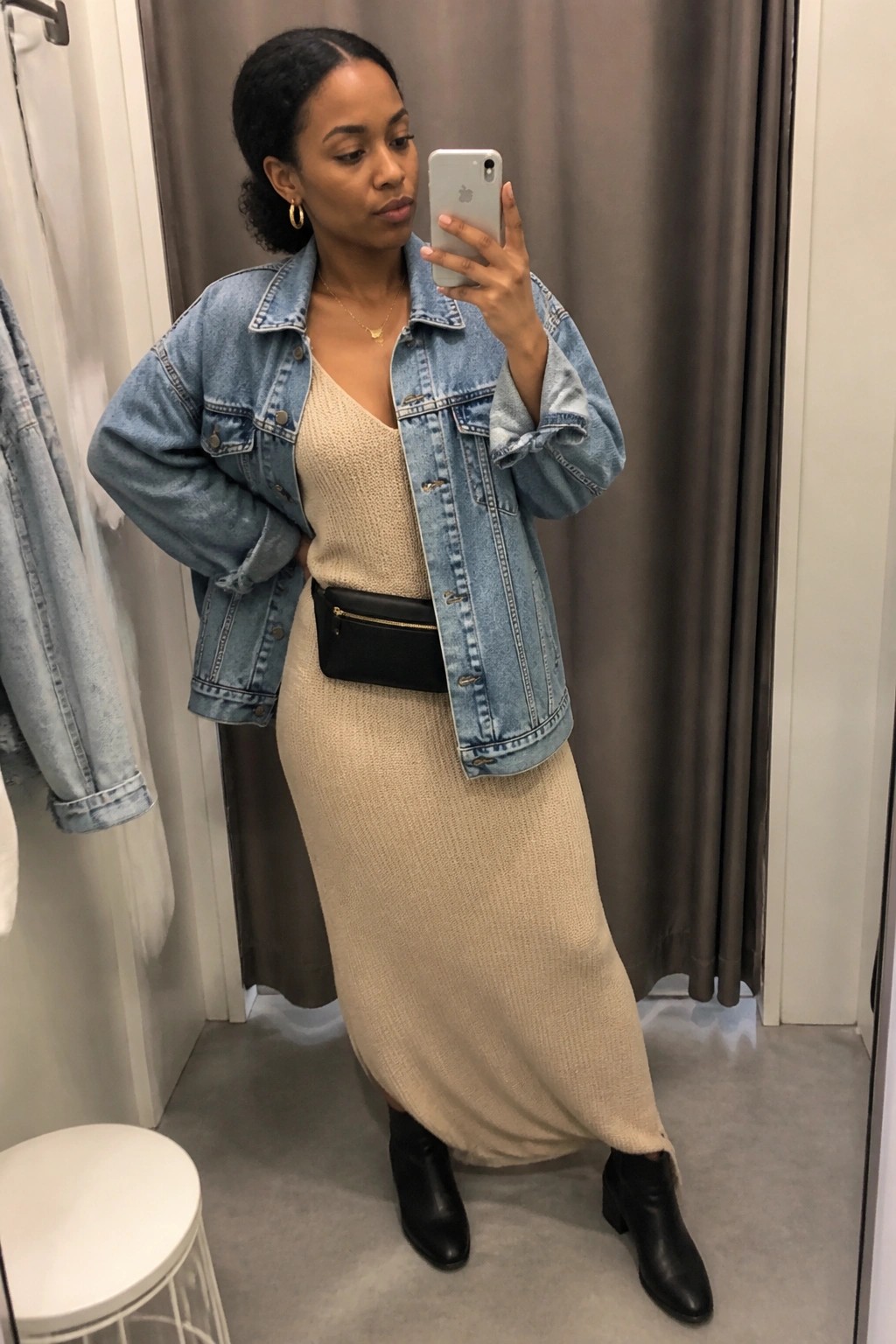 Woman in denim jacket over beige knit dress taking mirror selfie in fitting room
