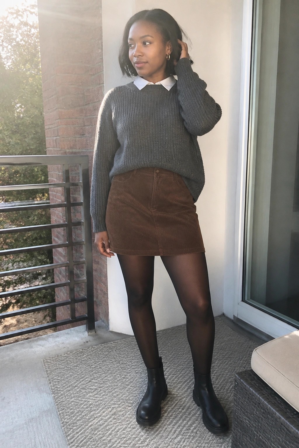 Woman in gray sweater over white collar, brown corduroy skirt, black tights and boots.