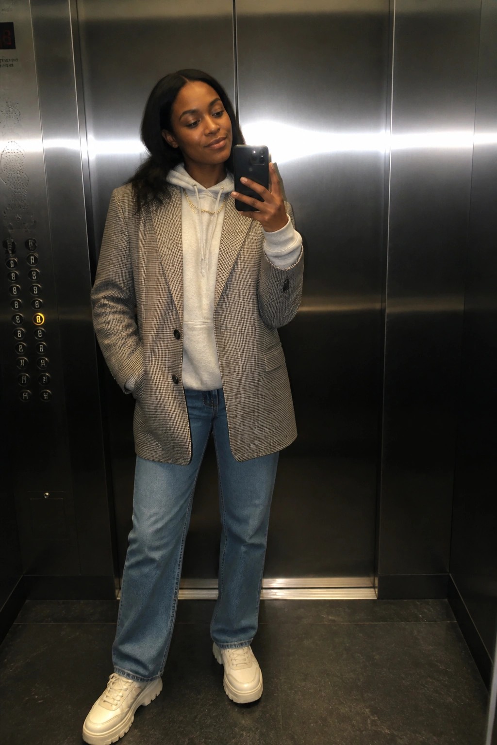 Woman in houndstooth blazer over hoodie taking selfie in elevator